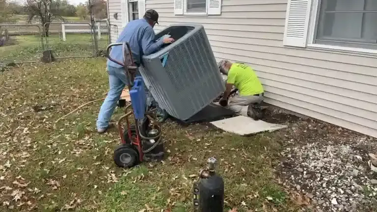 Two men installing a large, gray air conditioning unit near a house. One man uses a dolly.