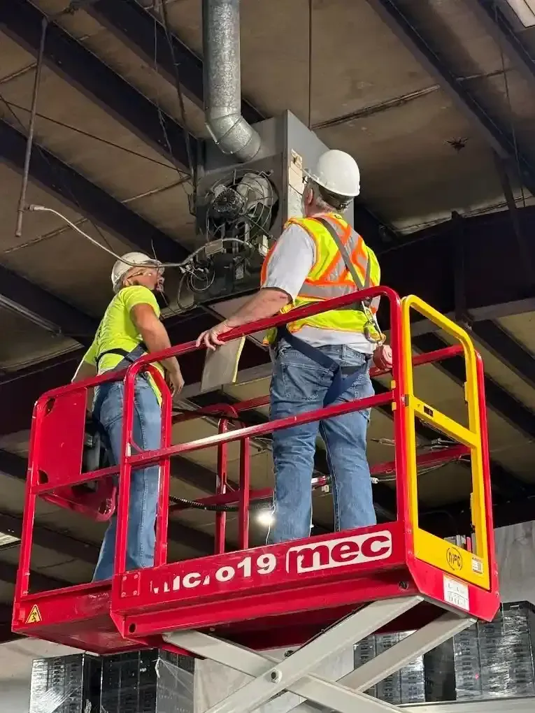Two workers on a red lift platform inspecting an industrial unit, wearing hard hats and safety vests.