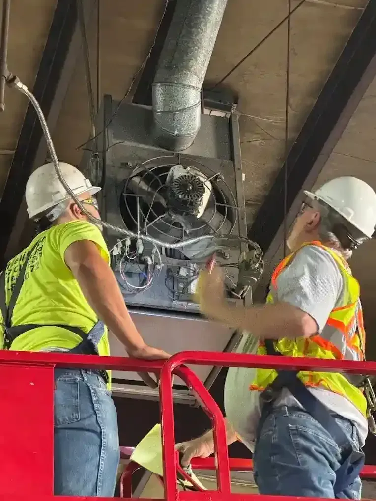 Two workers in hard hats and vests on a lift, inspecting HVAC unit.