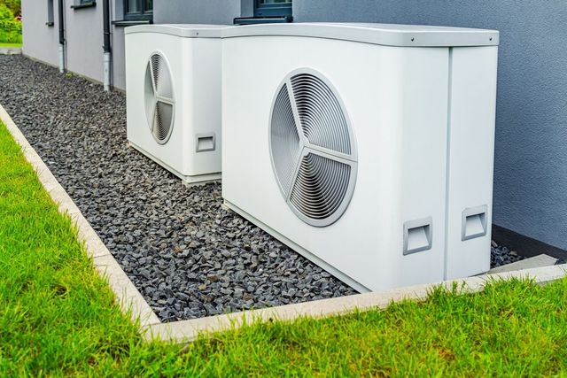 Two white heat pump units sit on gravel near a house, with green grass in foreground.