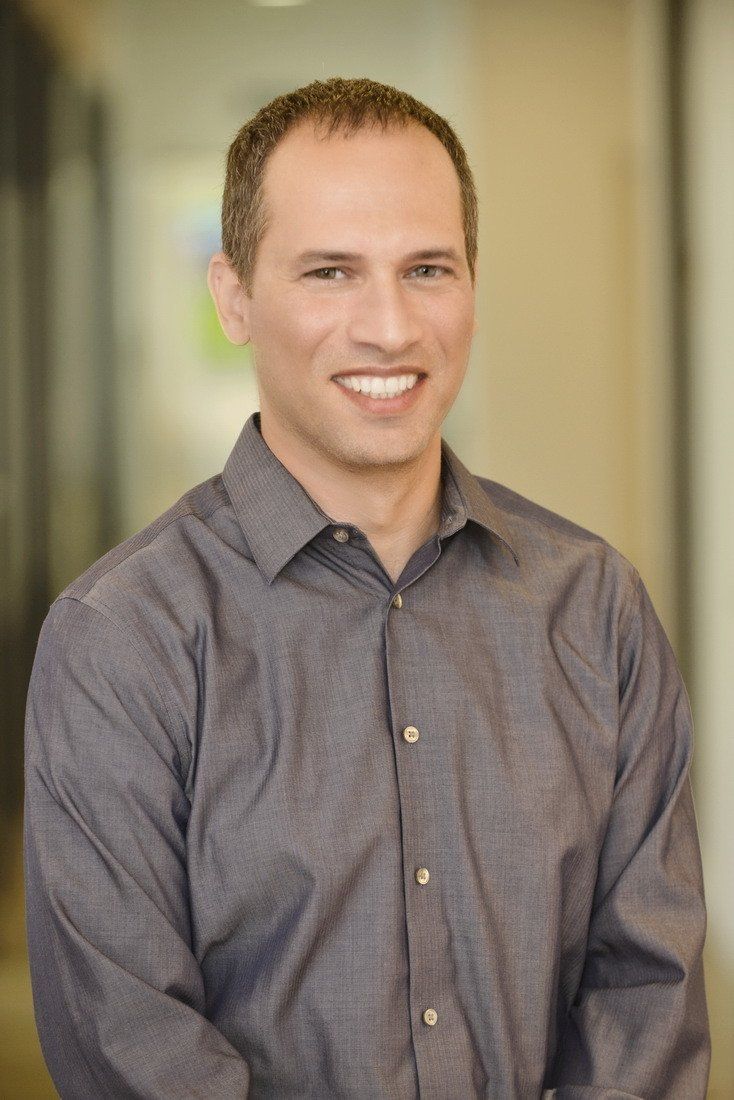 גבר בחולצה אפורה מחייך למצלמה.