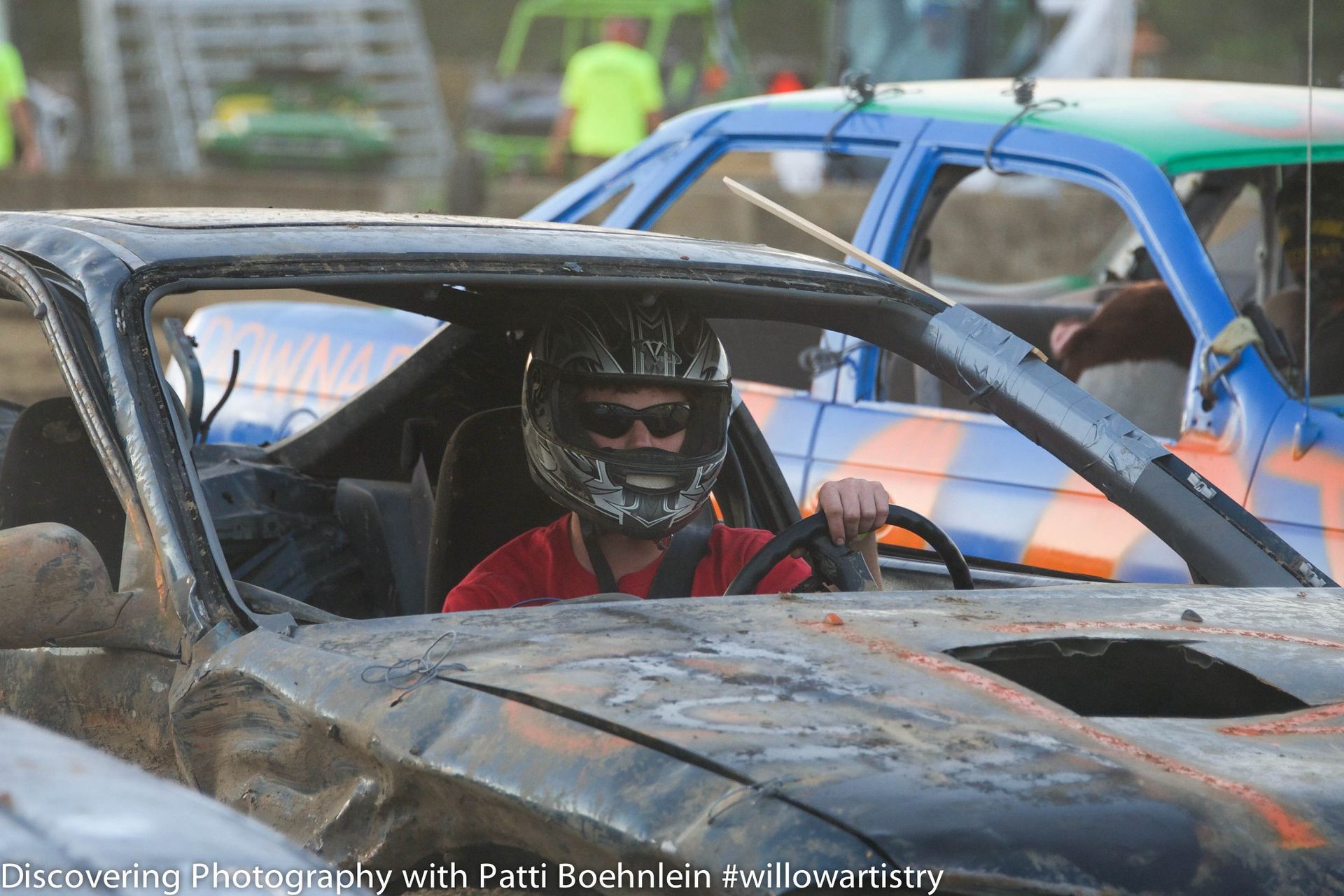 Driver in a black car, wearing a helmet, at a demolition derby. Another car is next to him.