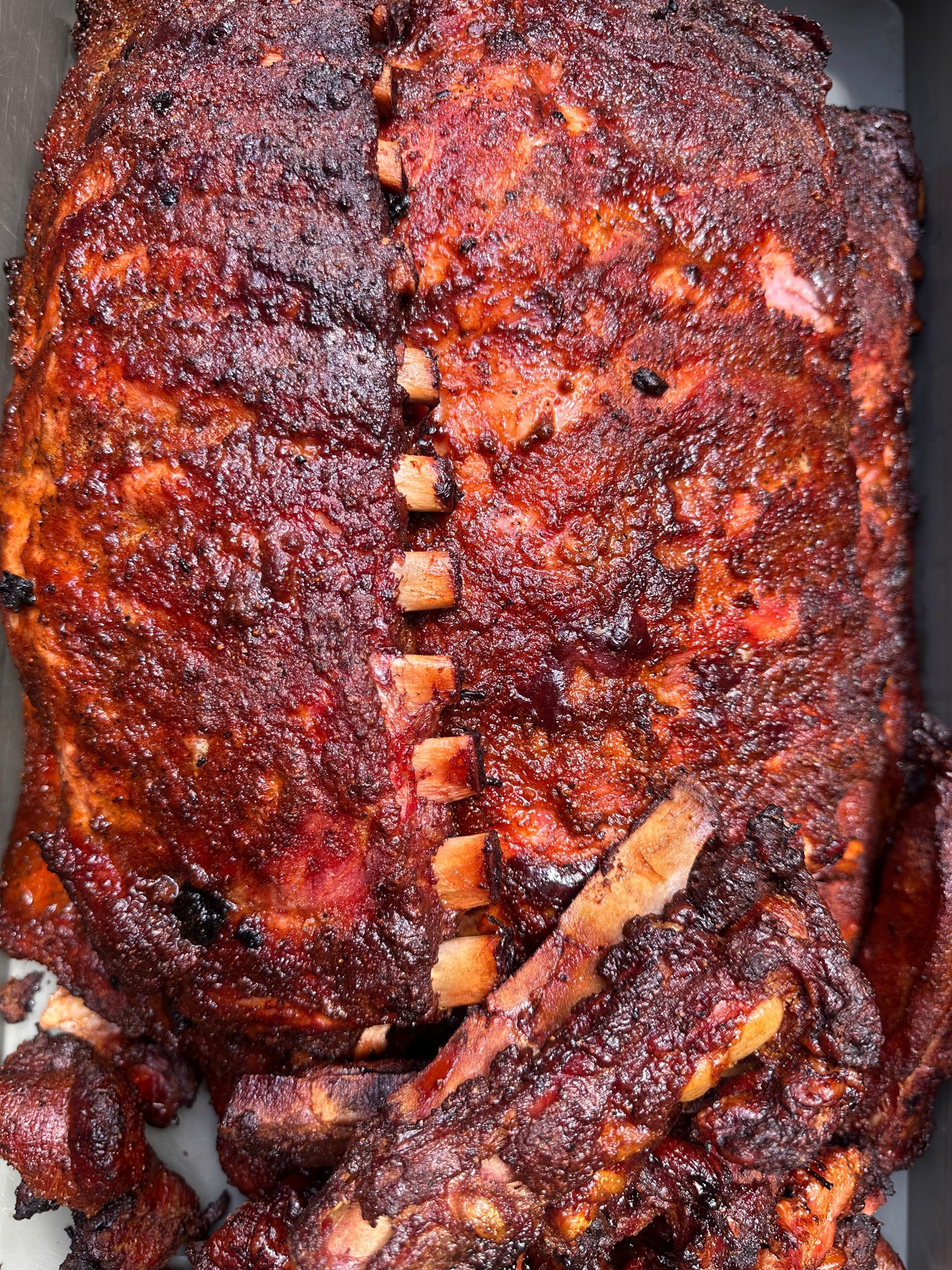 Close-up of glazed barbecue ribs on a tray, with dark charred edges and a glossy red-brown sauce.