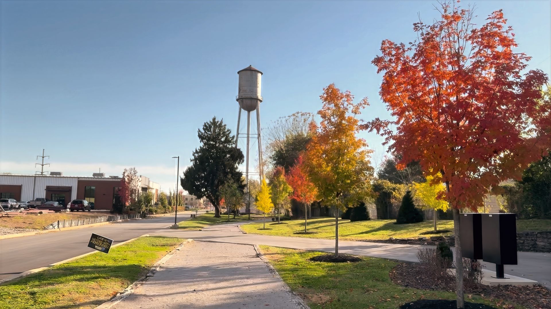 Park with pond, trees, and people. A modern building is in the background under a blue sky.