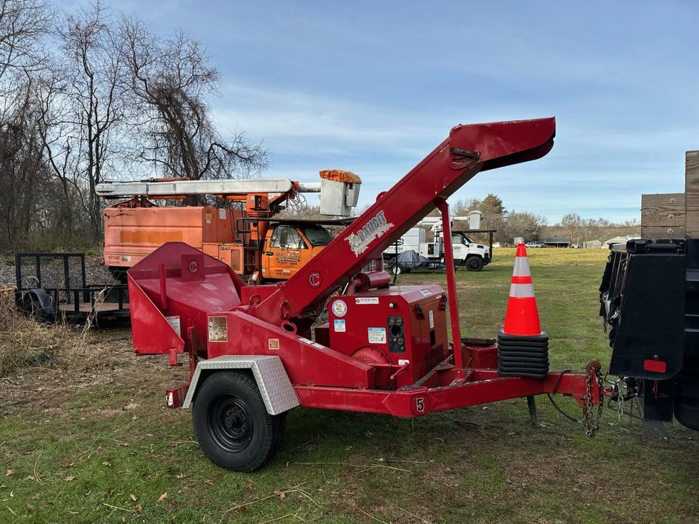 Red wood chipper on a trailer, parked on grass.  Orange cone in front, with utility trucks in the background.