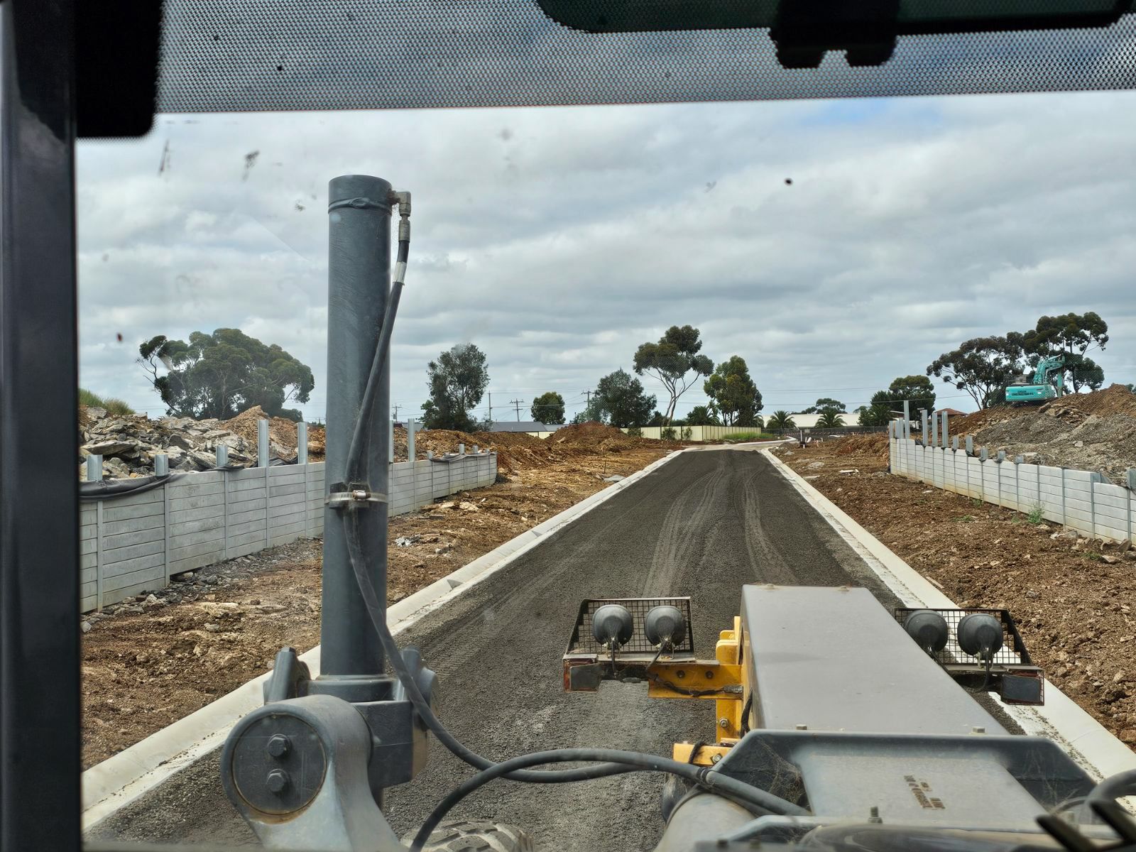 View from inside a grader operating on a road construction site; cloudy sky overhead.