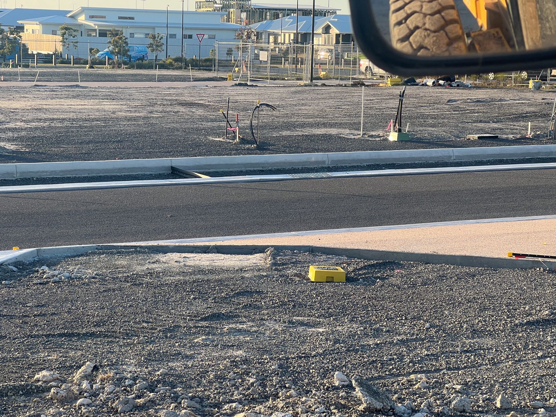 Construction site with a road, curb, gravel, and buildings in the background. A yellow box sits on the ground.