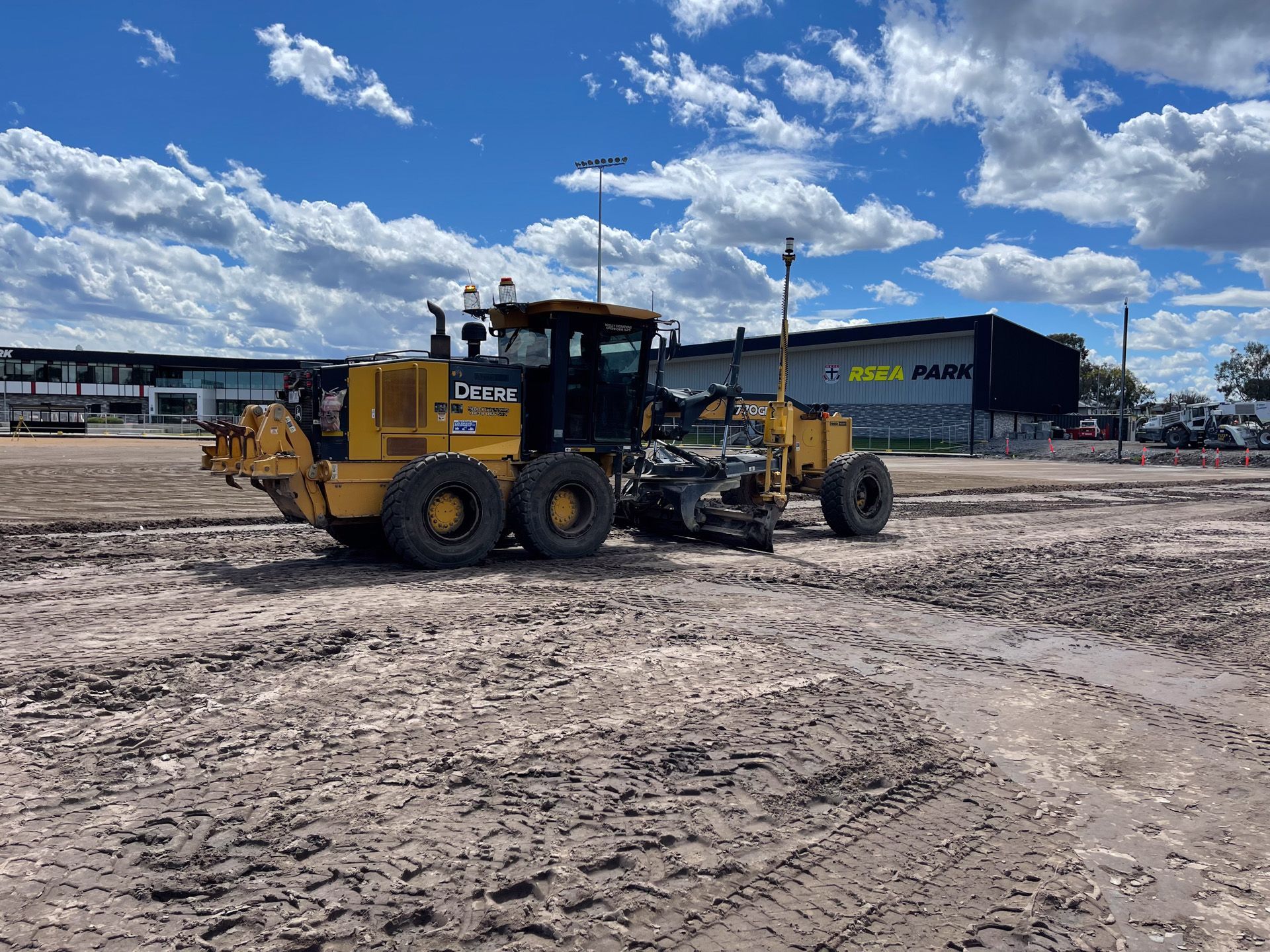 Yellow motor grader on dirt ground in front of a building under a cloudy sky.