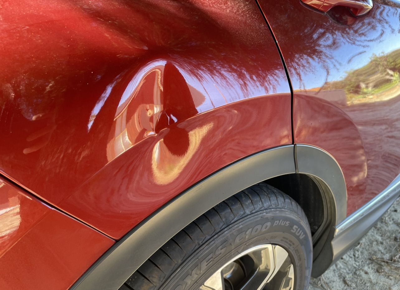 Red car with a large dent above the rear tire. Sunlight reflects off the damaged body.