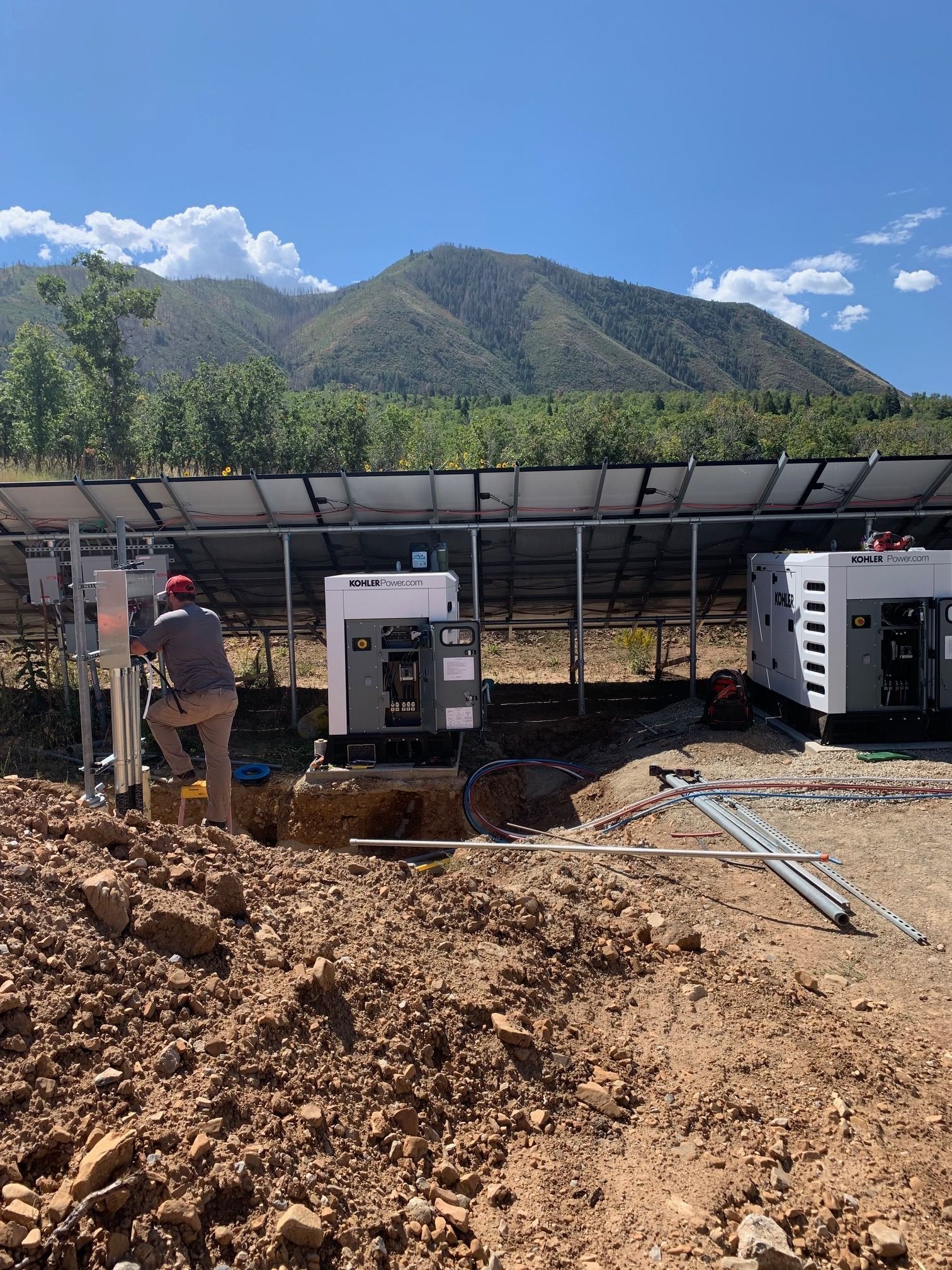 Construction worker near generators and solar panels on a hillside. Mountains and blue sky in background.