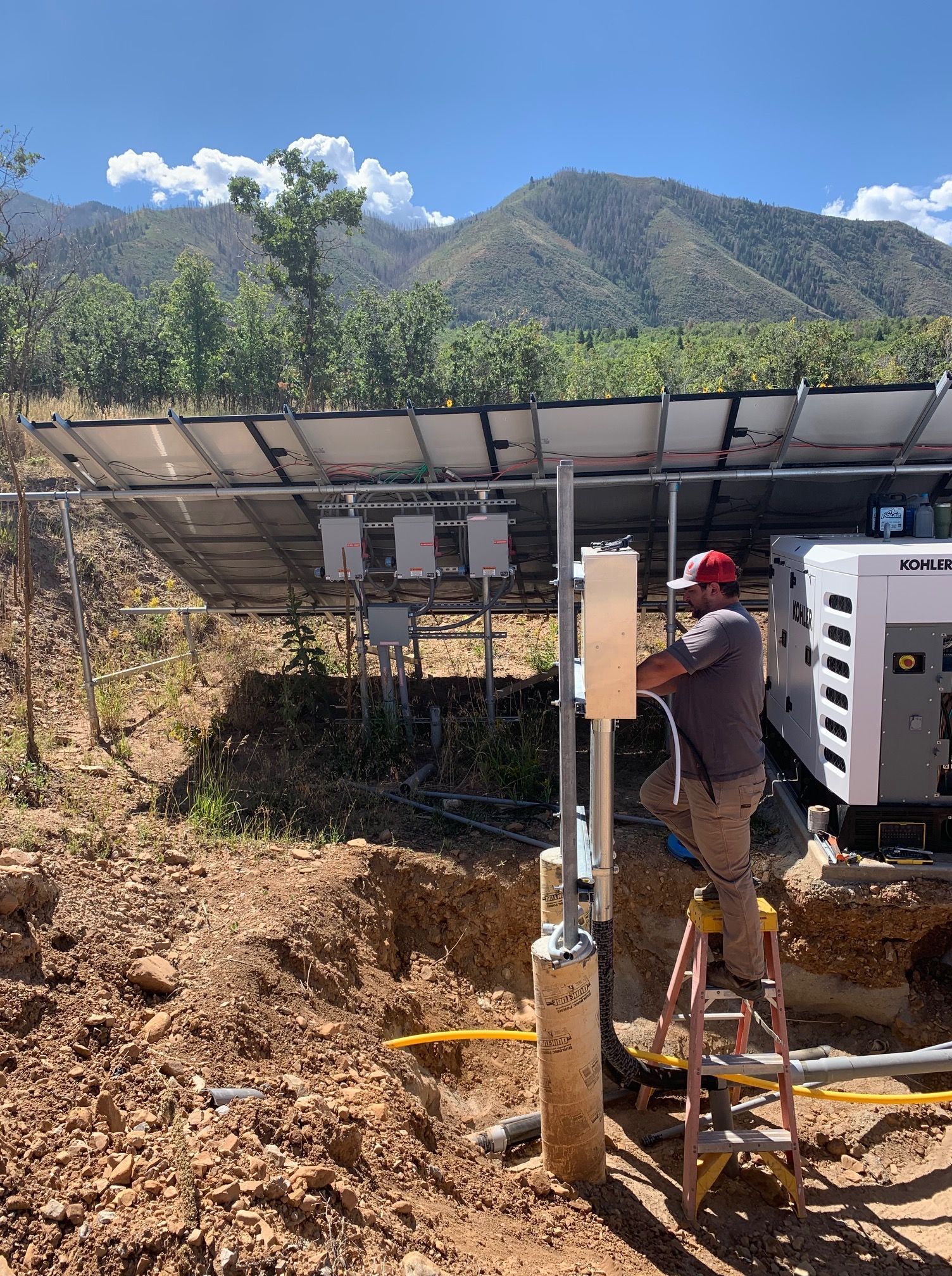 Man on ladder working on electrical box, solar panels in background. Mountain setting, sunny day.