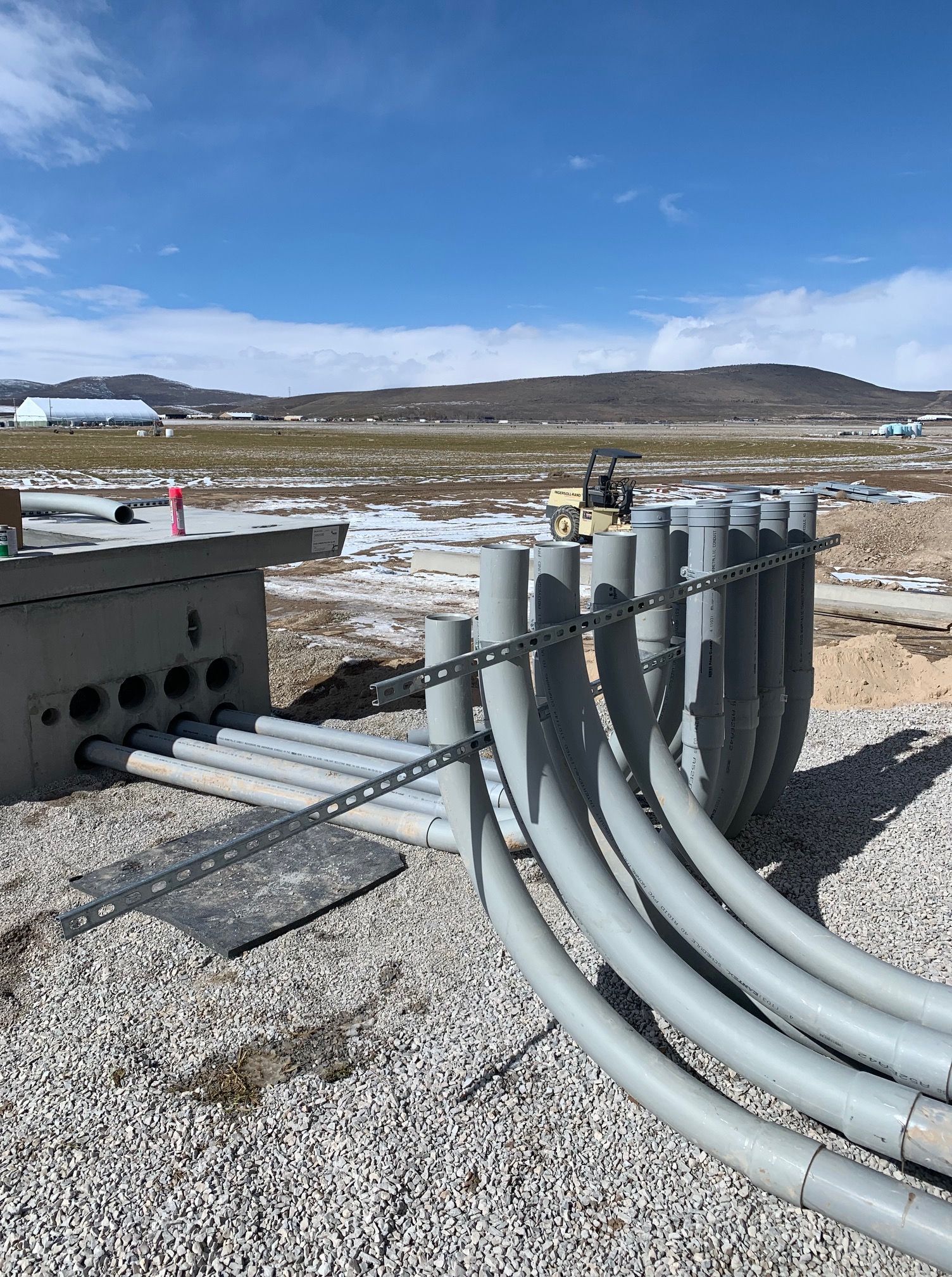 Gray conduit pipes exiting a concrete electrical box, construction site with blue sky and distant mountains.
