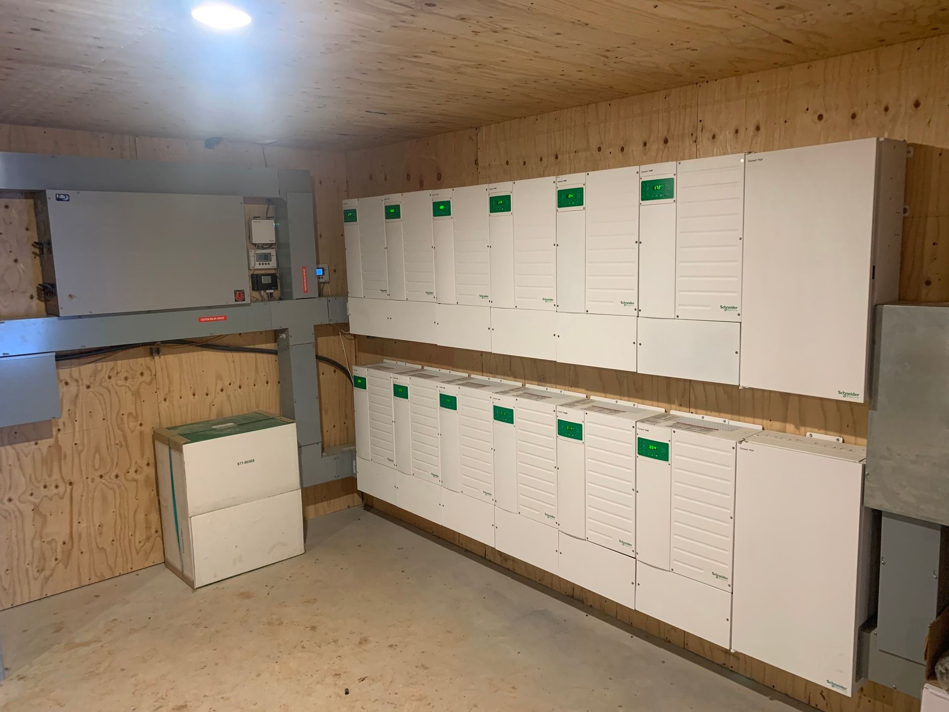 A room with solar panel inverters on wooden walls; a large electrical box is also present.