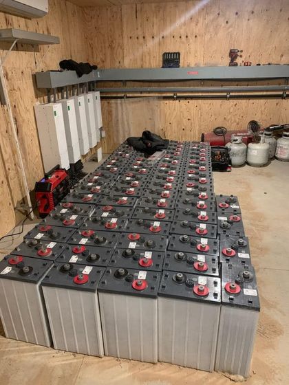 Rows of gray batteries in a wooden-walled room, possibly an off-grid power storage setup.