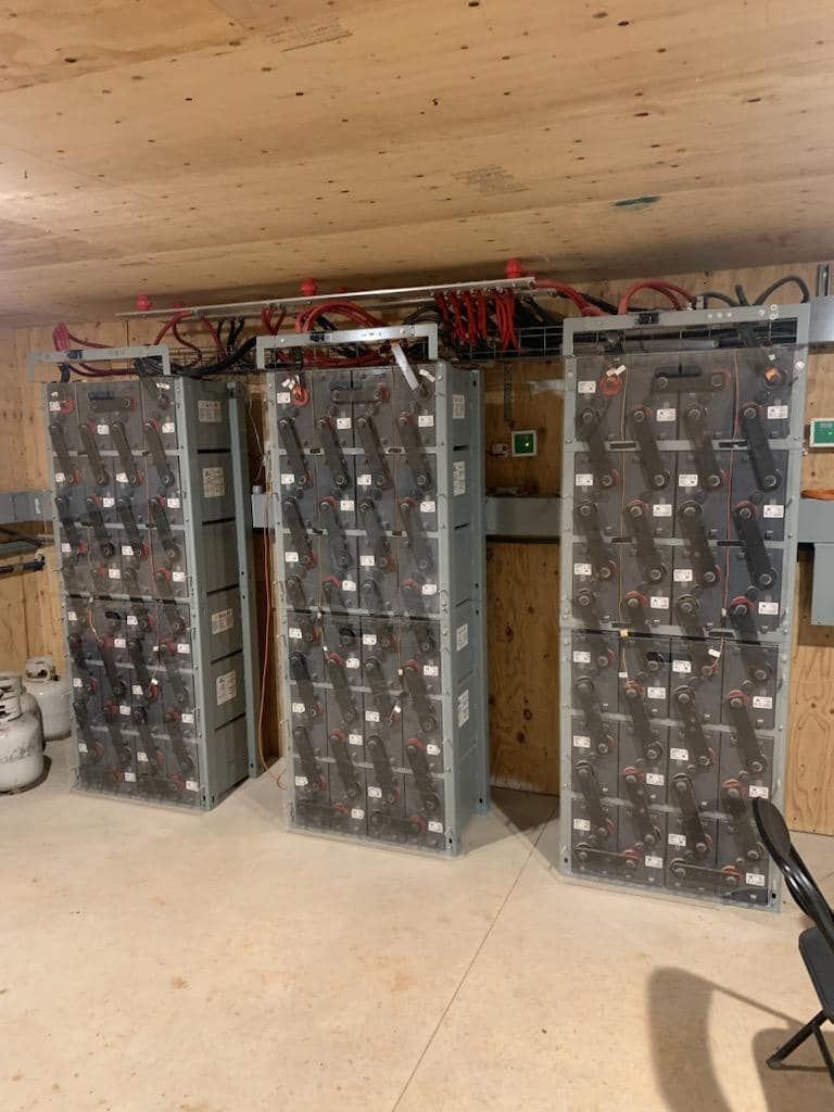 Three large battery stacks in a wooden-walled room, connected by overhead wiring.