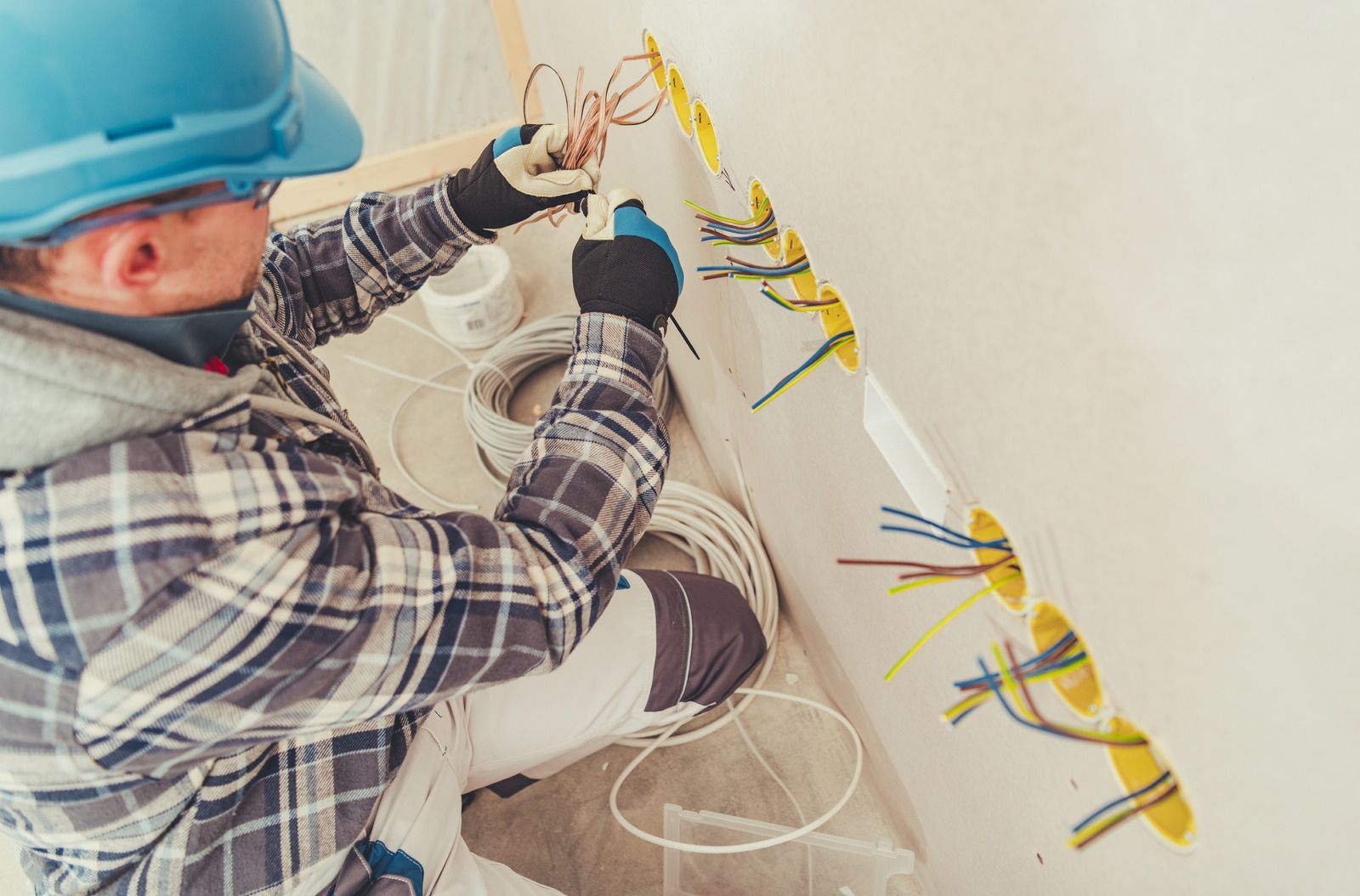 Electrician in blue hard hat and gloves working with wires in a wall.