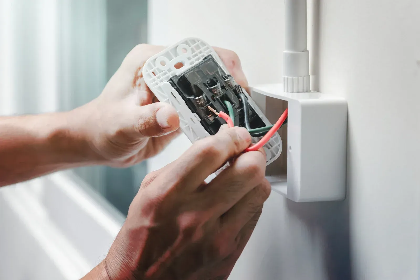 Person wiring an electrical outlet, hands working with red and green wires in a white box on a white wall.