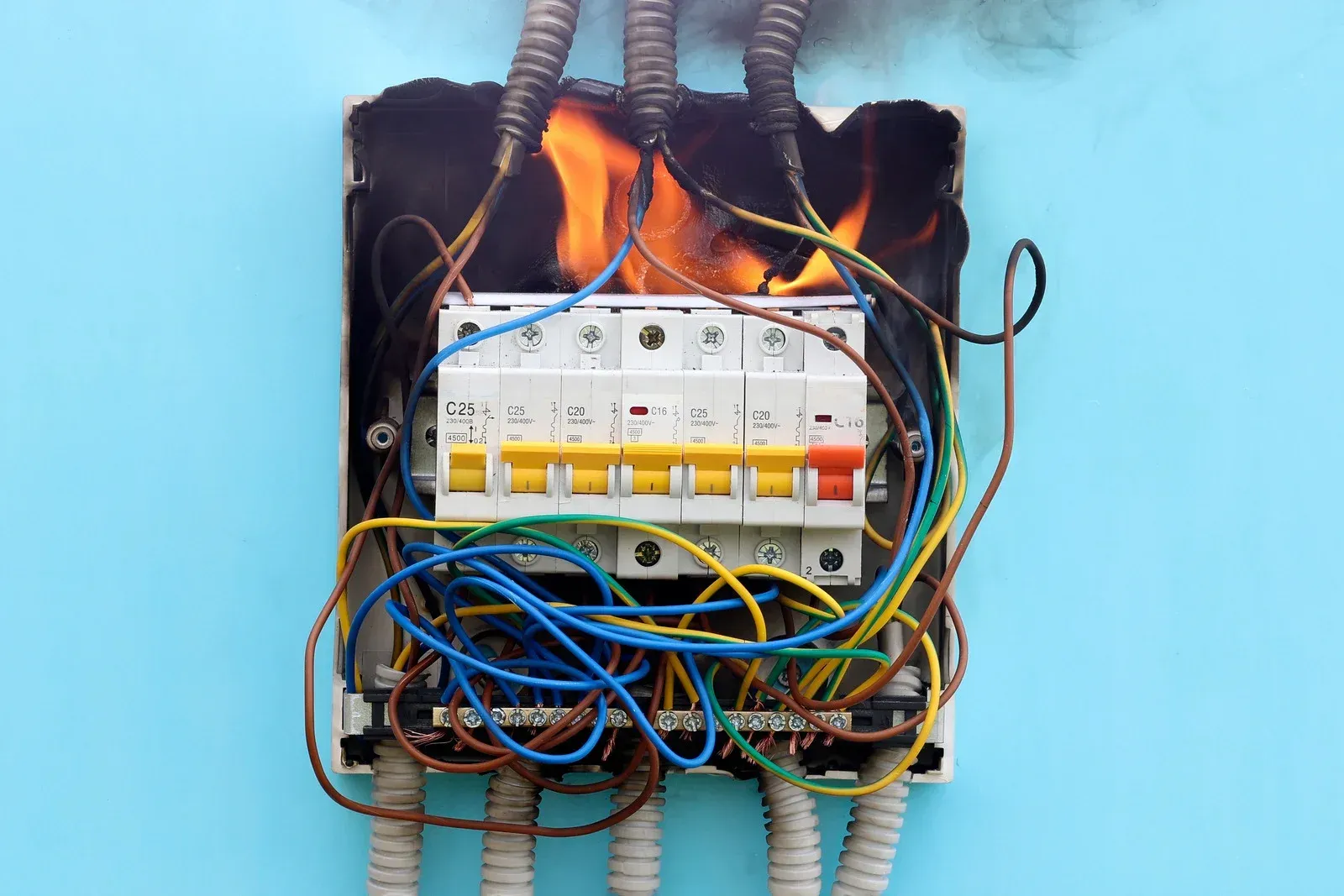 Electrical panel on fire, flames engulfing wires and circuit breakers, against a blue wall.