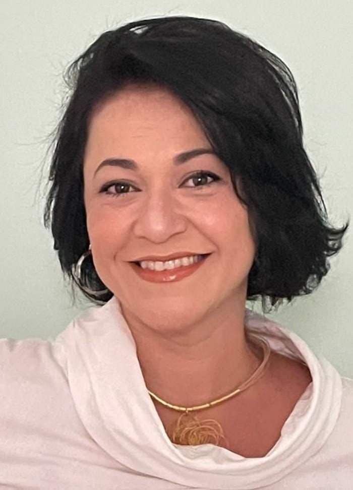 A woman wearing a white sweater and a gold necklace is smiling for the camera.