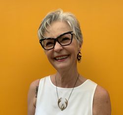 A woman wearing glasses and a necklace is smiling in front of a yellow wall.