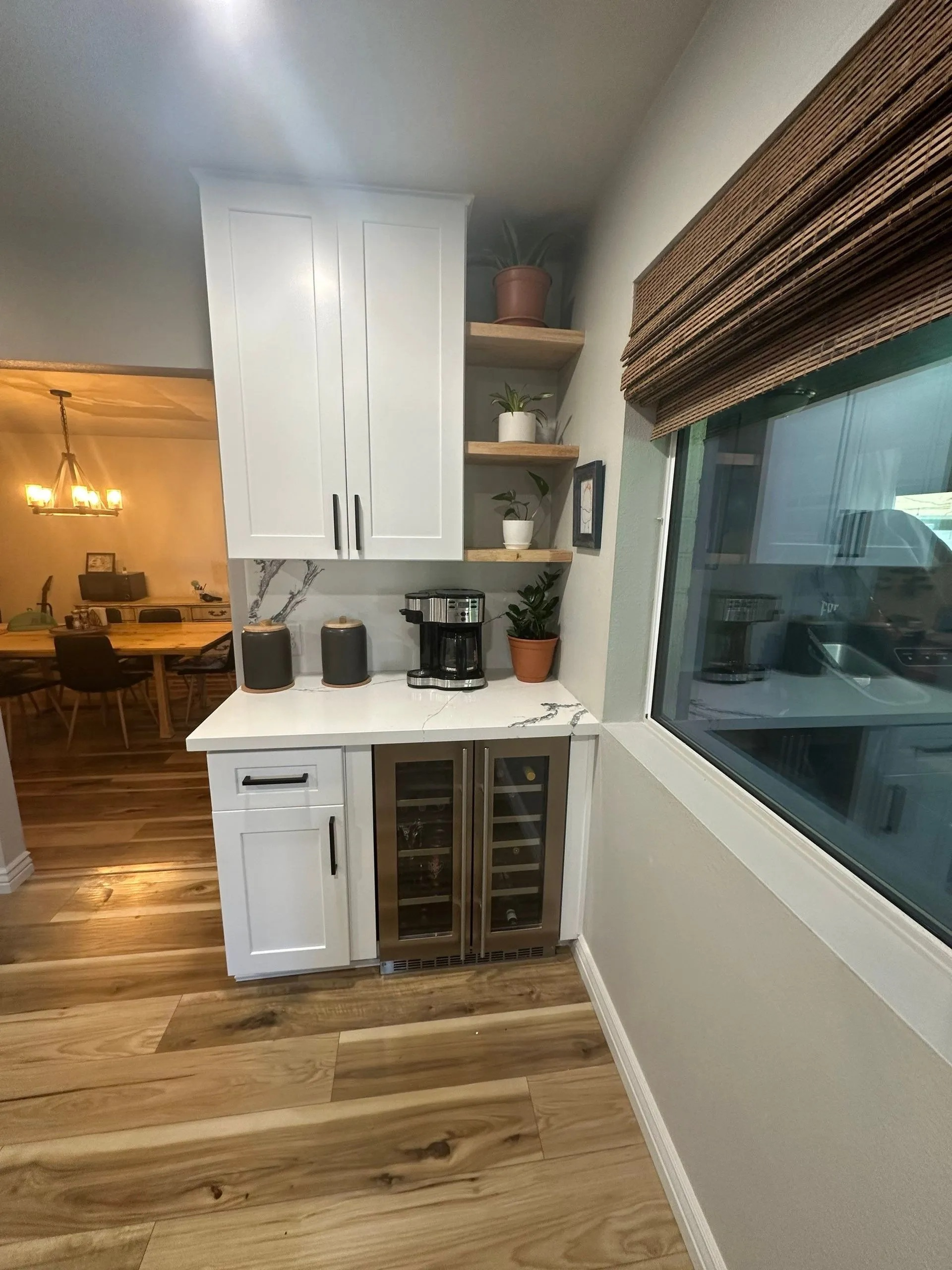 A kitchen with white cabinets , a wine cooler , a coffee maker , and a window.