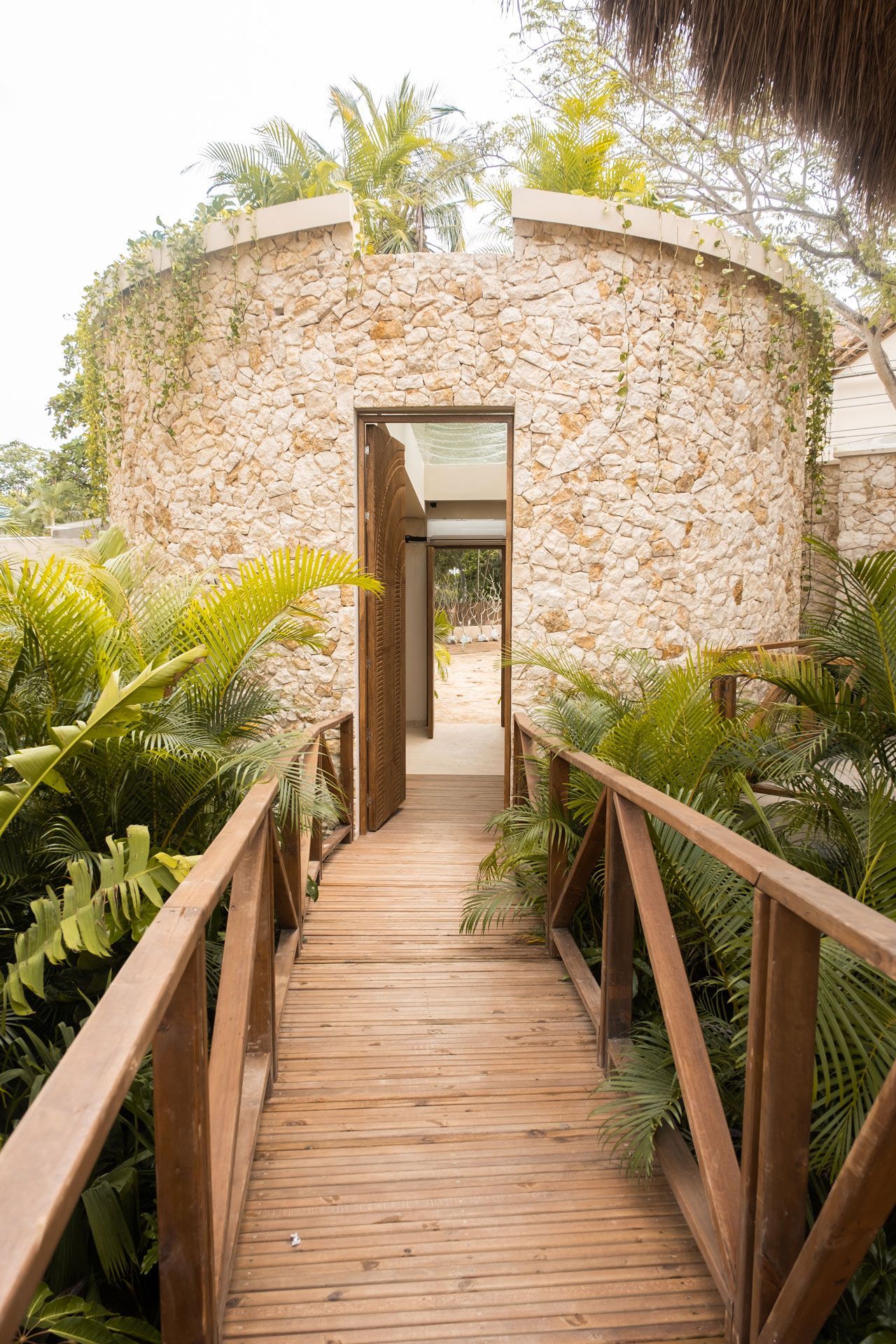 Entrada de Aimarawa en San Antero con pasarela de madera, plantas tropicales y fachada de piedra.