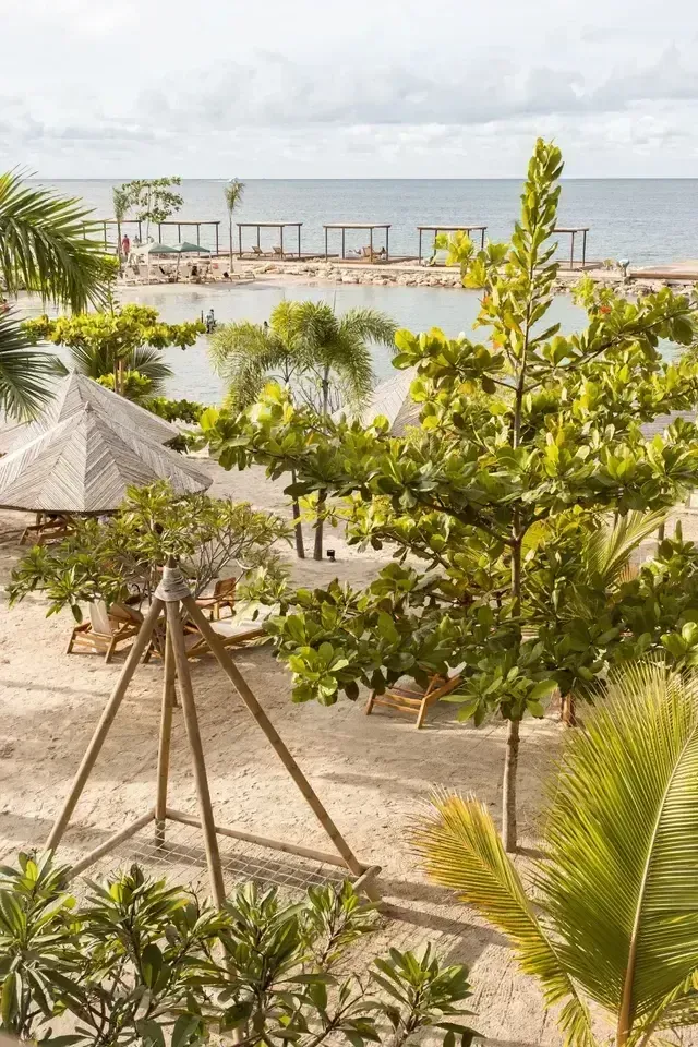 Vista desde el Hotel Aimarawa hacia la playa privada con arena clara, palmeras y sombrillas de palma, junto a una piscina natural frente al mar Caribe.