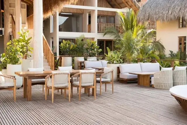 Terraza exterior del Hotel Aimarawa con mesas de madera, sofás lounge y sillas artesanales, rodeada de vegetación tropical y arquitectura en palma.