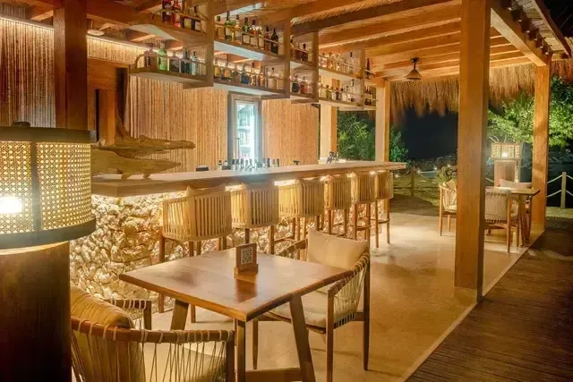 Bar Ara Macao del Hotel Aimarawa con barra en piedra y madera, sillas altas artesanales y estanterías con licores, iluminado en un ambiente cálido y tropical.