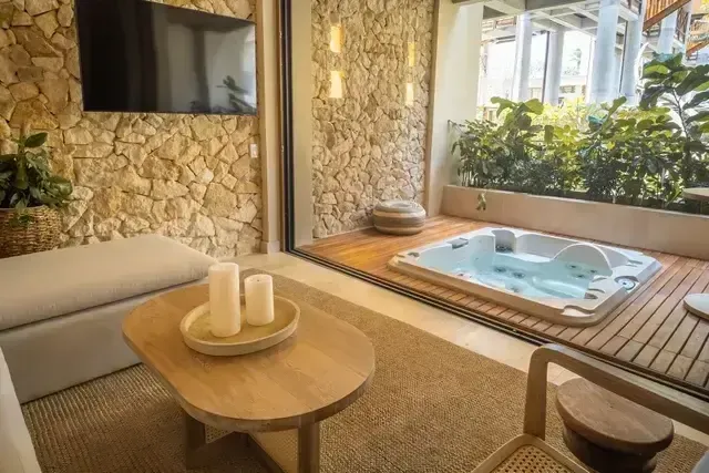 Suite del Hotel Aimarawa con diseño natural, pared en piedra, sala acogedora con mesa de madera y velas decorativas, junto a terraza privada con jacuzzi rodeado de vegetación.