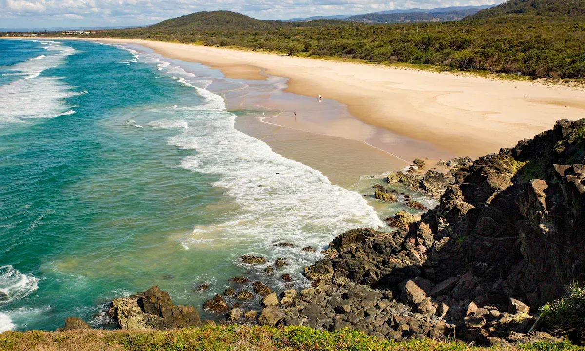 Ocean waves crashing on a sandy beach, with a rocky foreground and forested hills in the background. — North Coast Stock Feeds in Wardell, NSW