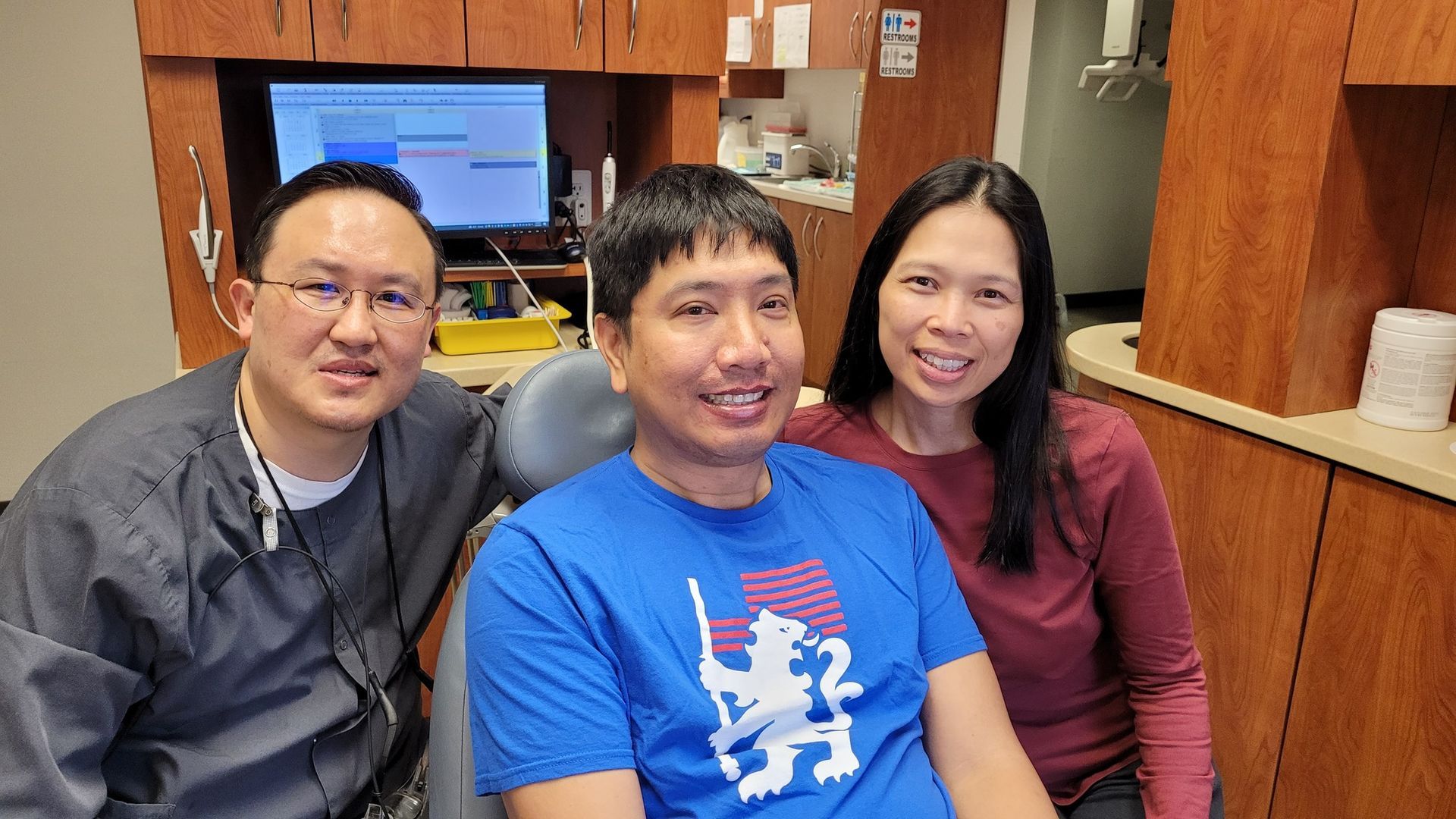 A man smiles in a dental chair with two people, one in scrubs, another in a maroon shirt, in a dental office.
