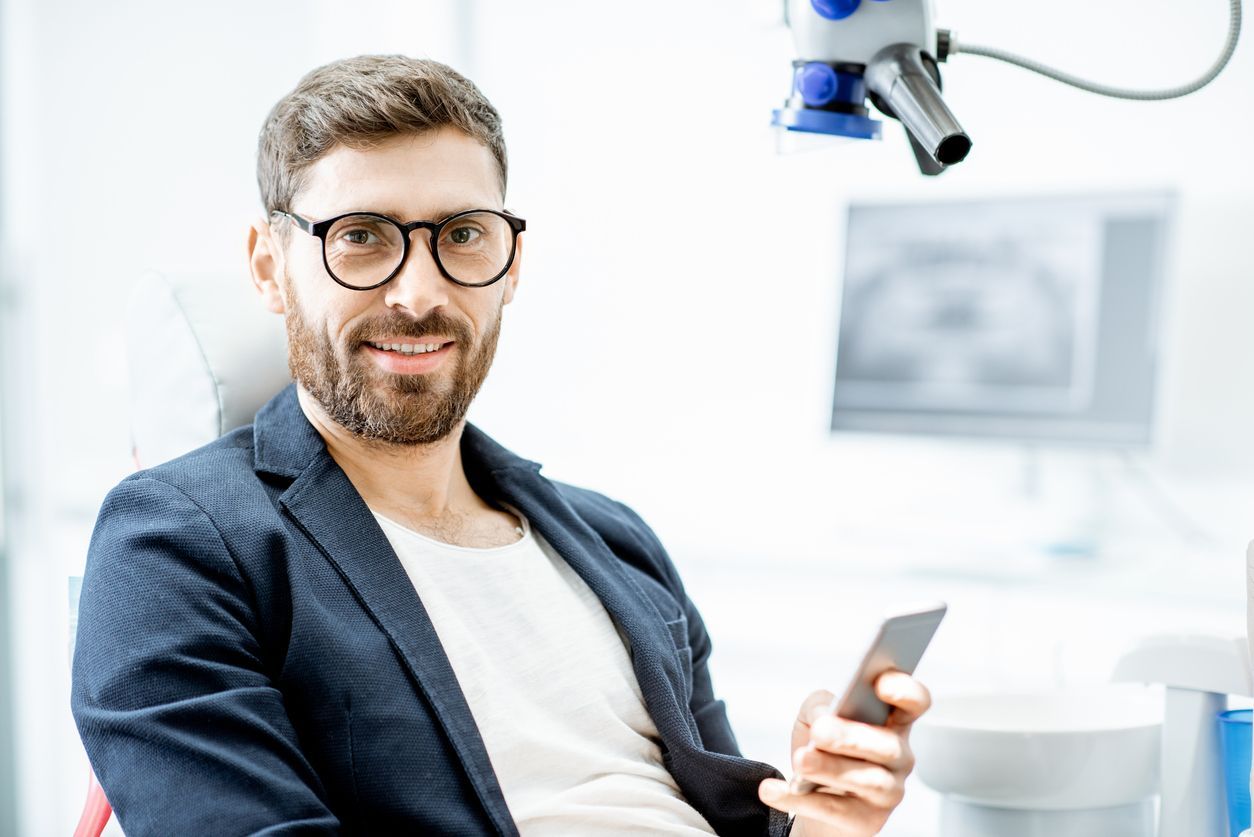 Man in glasses and blazer smiles in a dental chair, holding a phone. Dental office setting.