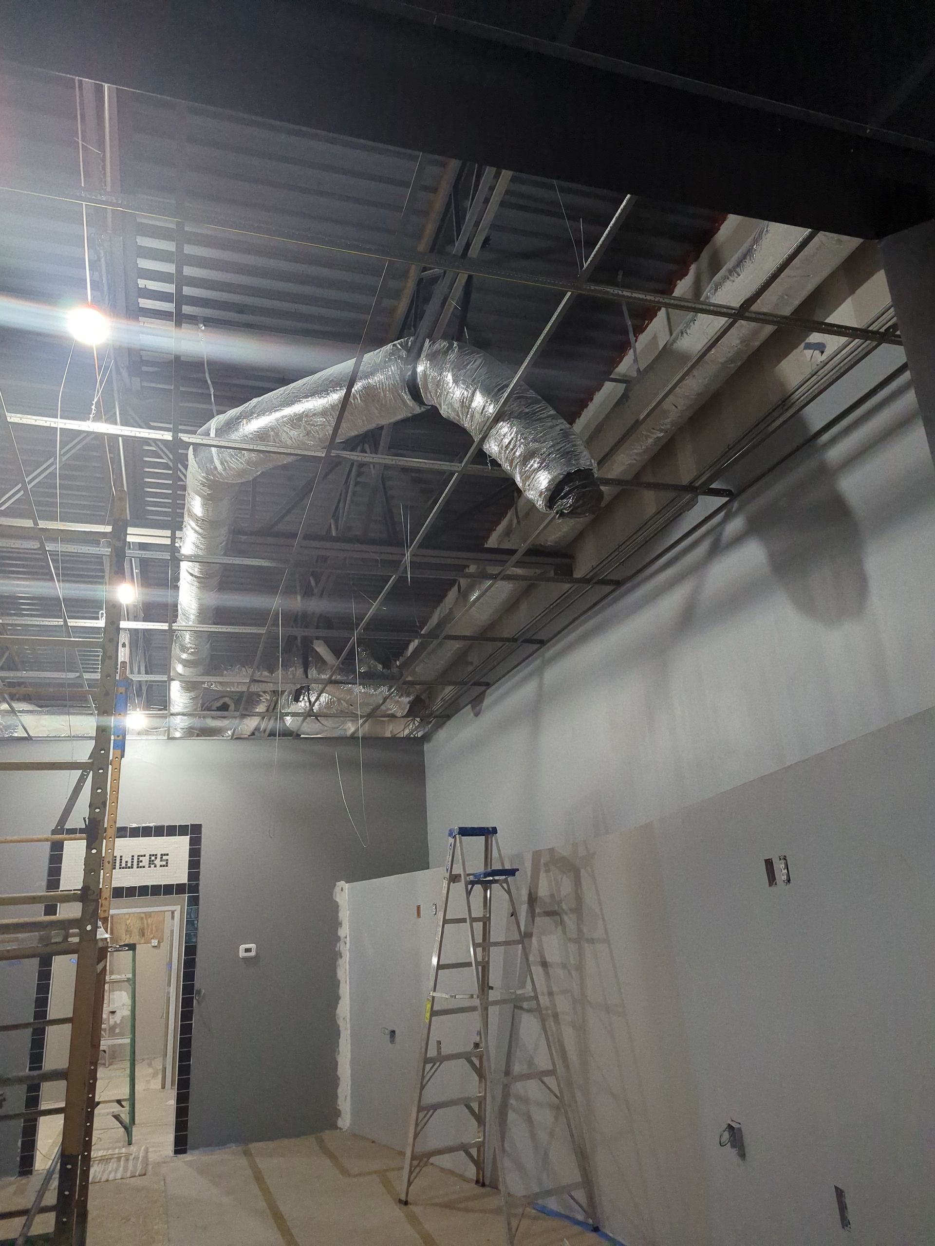 An interior construction site showing an exposed ceiling with flexible ductwork and a metal ladder against a grey wall.