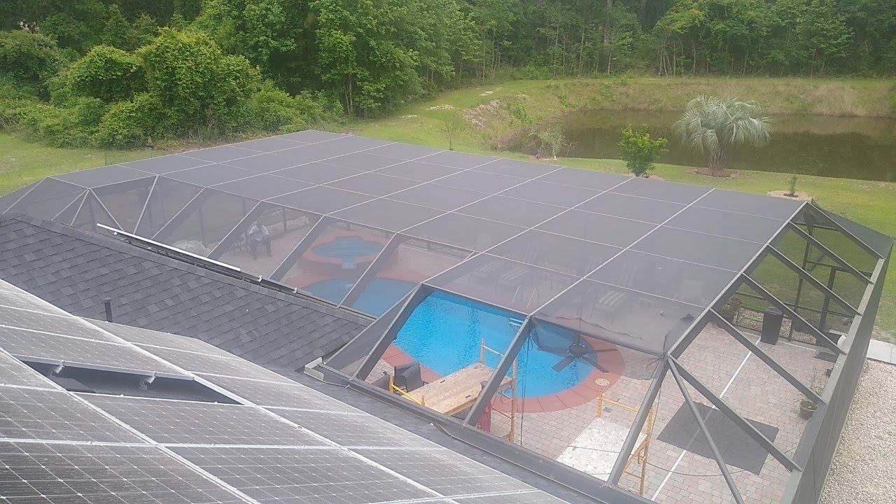 An aerial view of a house roof with solar panels next to a screened-in swimming pool enclosure in a grassy backyard.