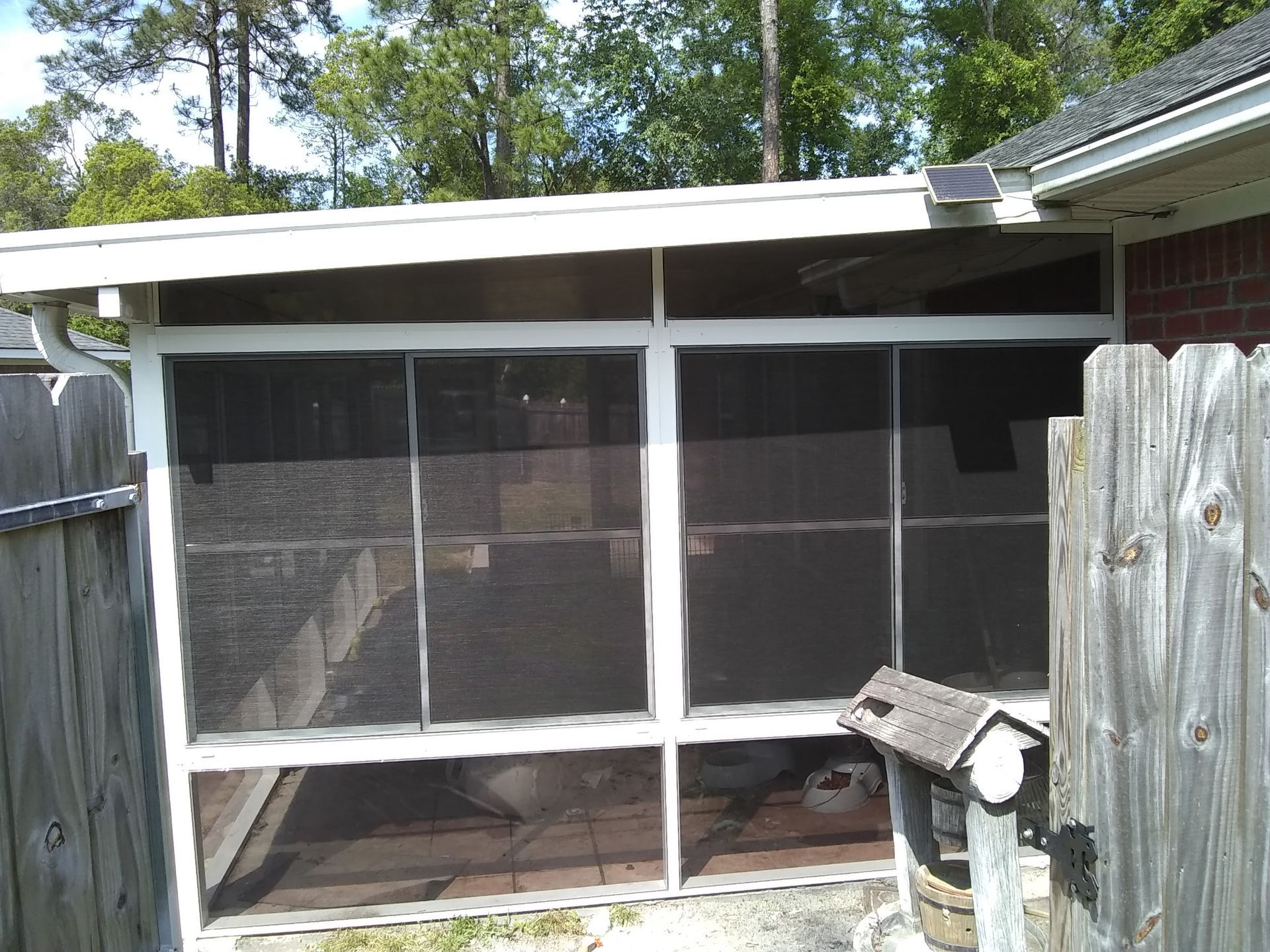 A white, screened-in patio area attached to a brick house, flanked by wooden fences on both sides.