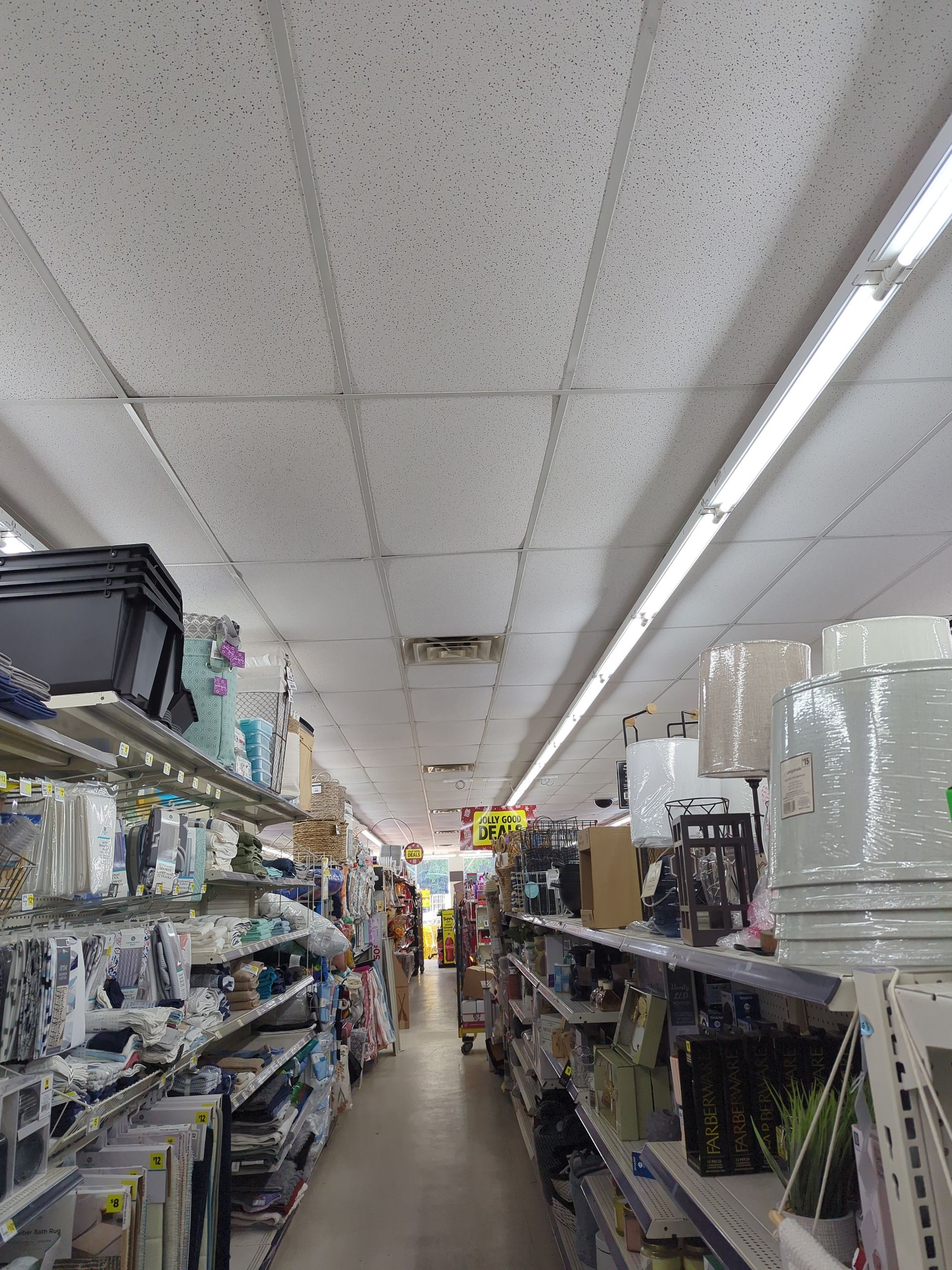 An aisle in a retail store filled with various home goods on shelves, extending towards a brightly lit storefront.