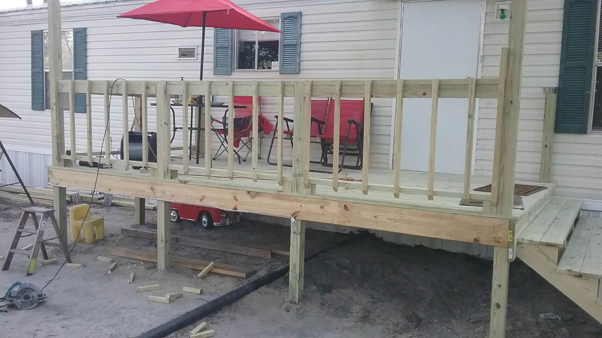 A partially constructed wooden deck with railings attached to the front of a light-colored mobile home with a red umbrella.
