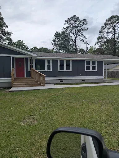 A single-story, dark gray house with a red front door, wood steps, and a concrete walkway in a green, grassy yard.