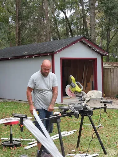 A person operating a miter saw on a stand outside in front of a gray shed with red trim.