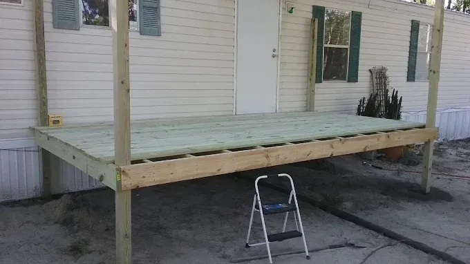 A wooden deck frame under construction against the side of a manufactured home, with a small stepladder underneath.