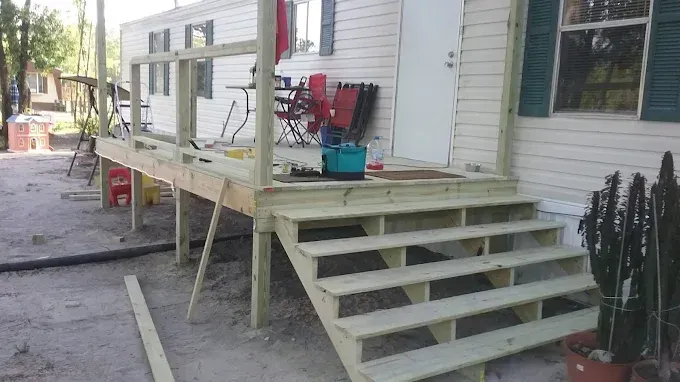 A wooden porch and staircase under construction attached to the side of a light-colored mobile home.