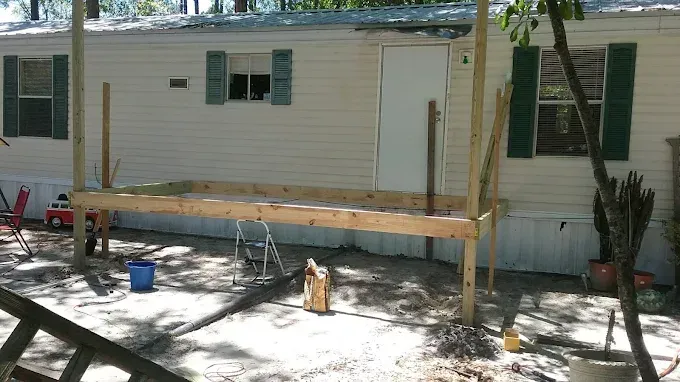 A wooden deck frame under construction in front of a mobile home with light siding and green shutters.