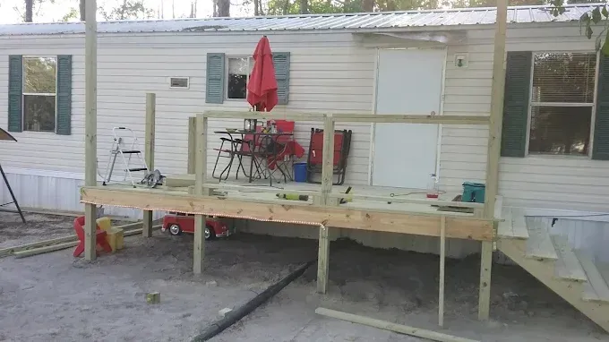 A partially constructed wooden deck with railings attached to the front of a light-colored mobile home.