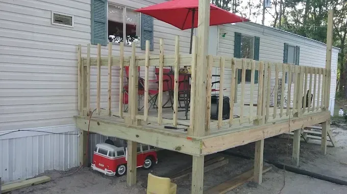A new wooden deck attached to a light-colored mobile home, featuring a red umbrella and a toy vehicle parked underneath.