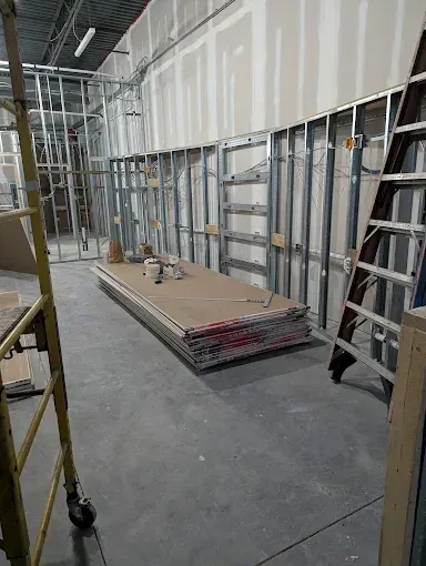 Construction site interior with a stack of drywall sheets, metal wall framing, and step ladders on a concrete floor.
