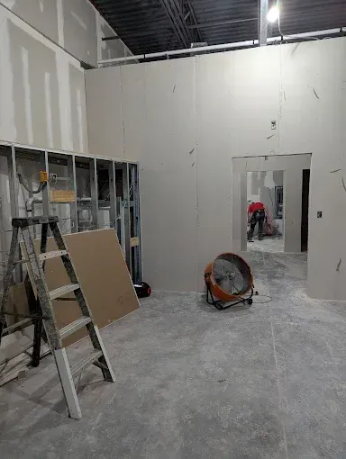 A construction site interior featuring unfinished drywall, a metal ladder, and a floor fan with a worker visible in a doorway.