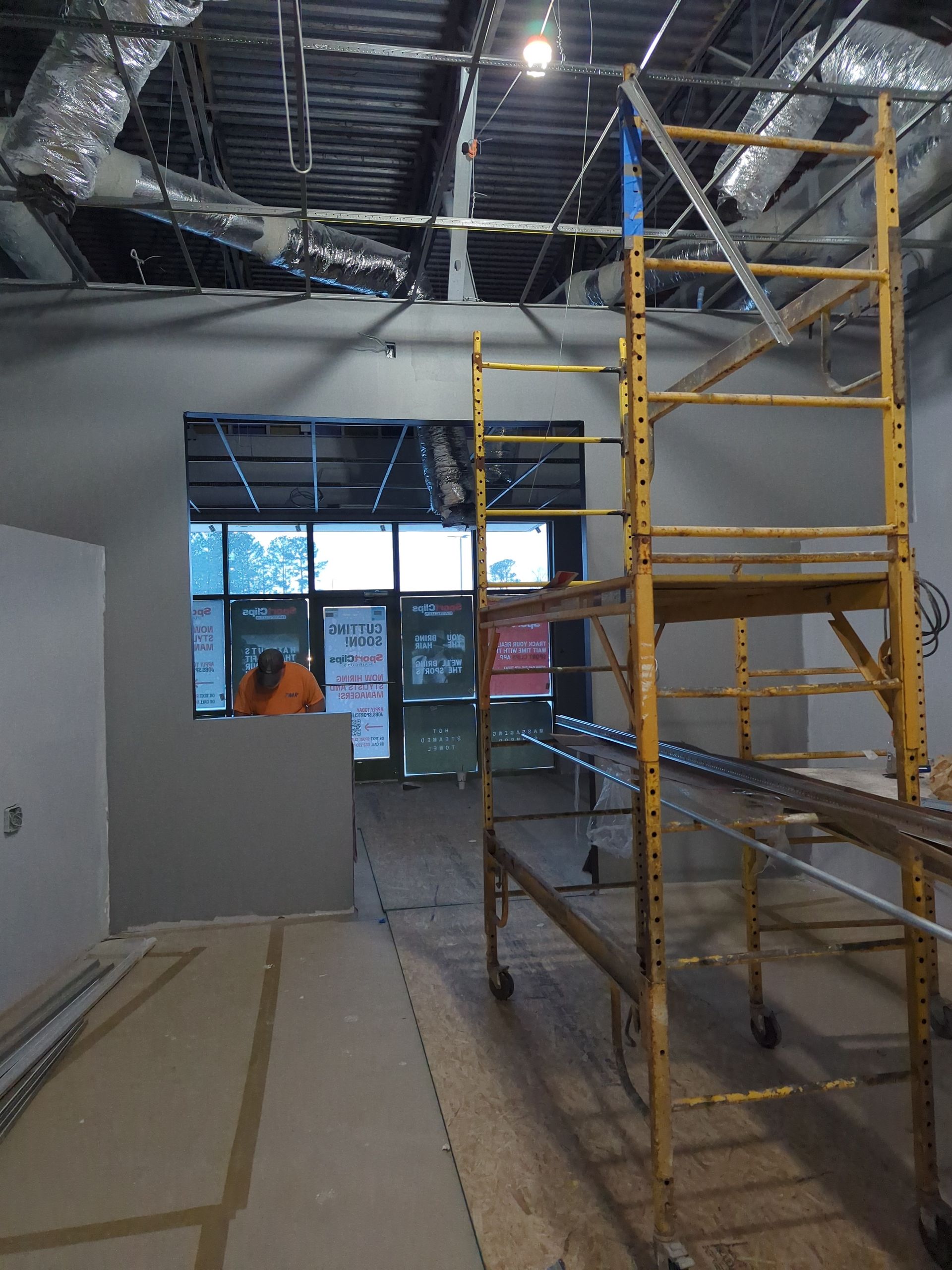 A person in an orange shirt works in a room under construction with a large yellow scaffold, grey walls, and exposed pipes.