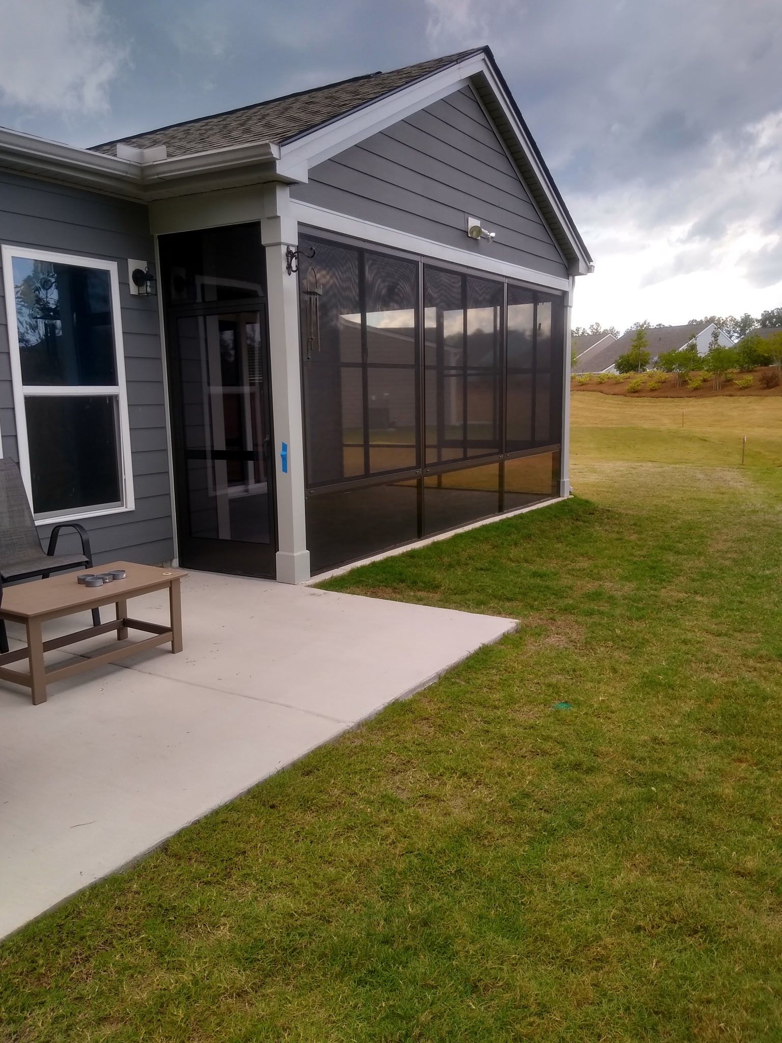 A gray house with a screened-in porch and patio area, overlooking a grassy yard under a cloudy sky.