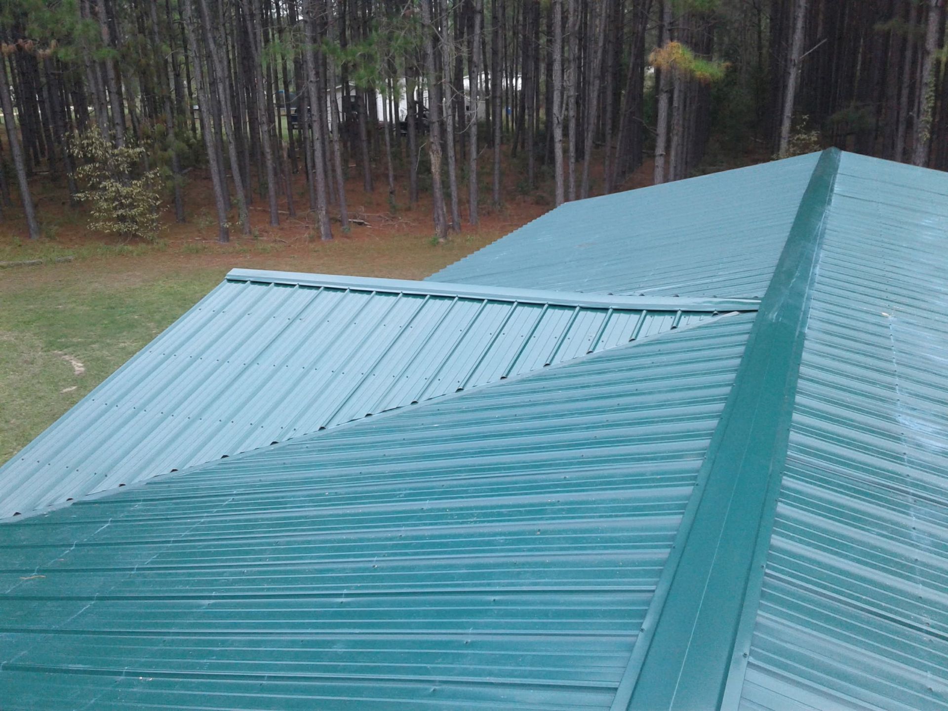 Green metal roof with visible ridges and seams, showing a valley where two roof sections meet near a forested backyard.